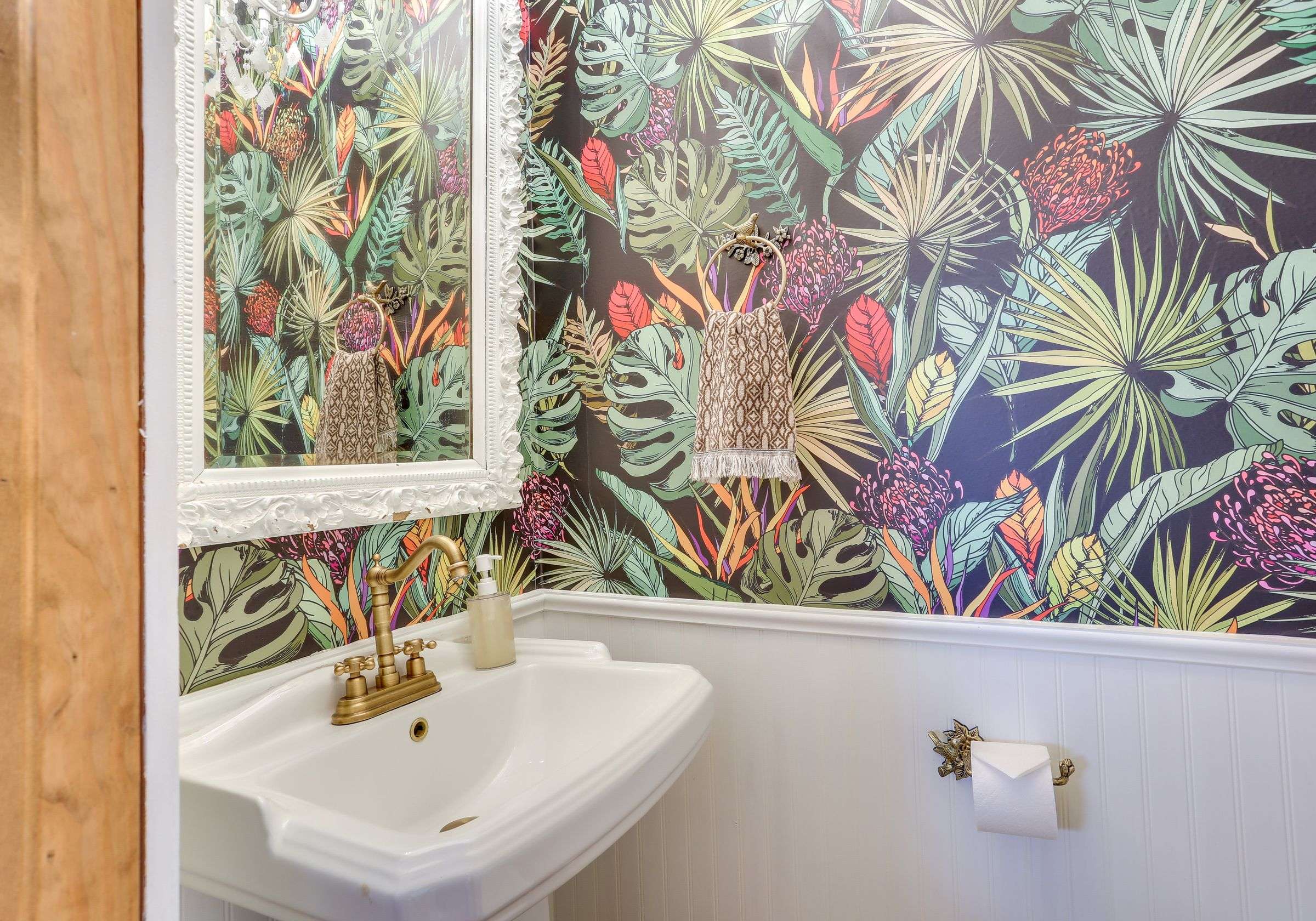 The half bath with bold botanical wallpaper, a pedestal sink and an ornate white mirror.
