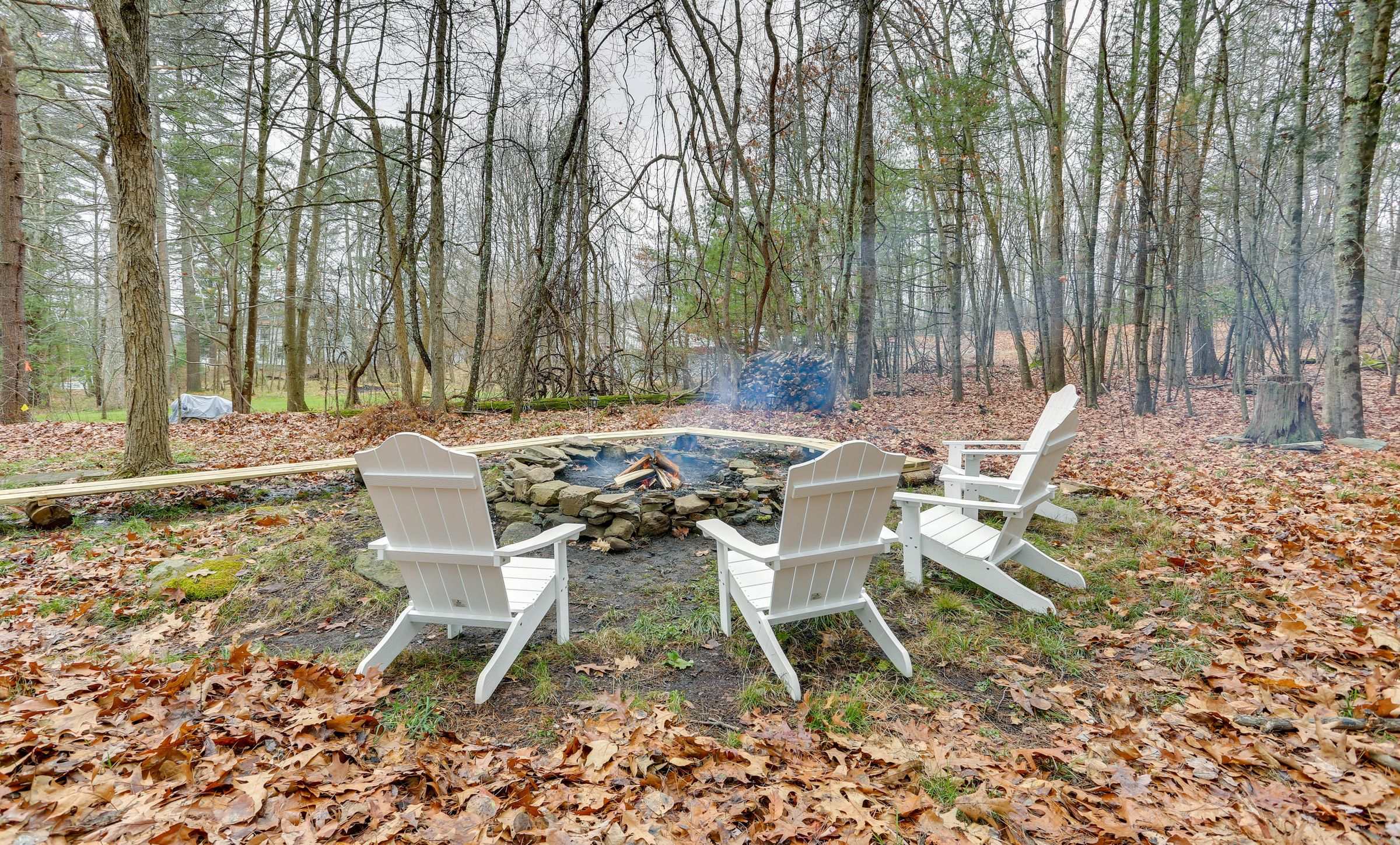 The stone fire pit at the edge of the woods, white chairs on a bed of fallen leaves.