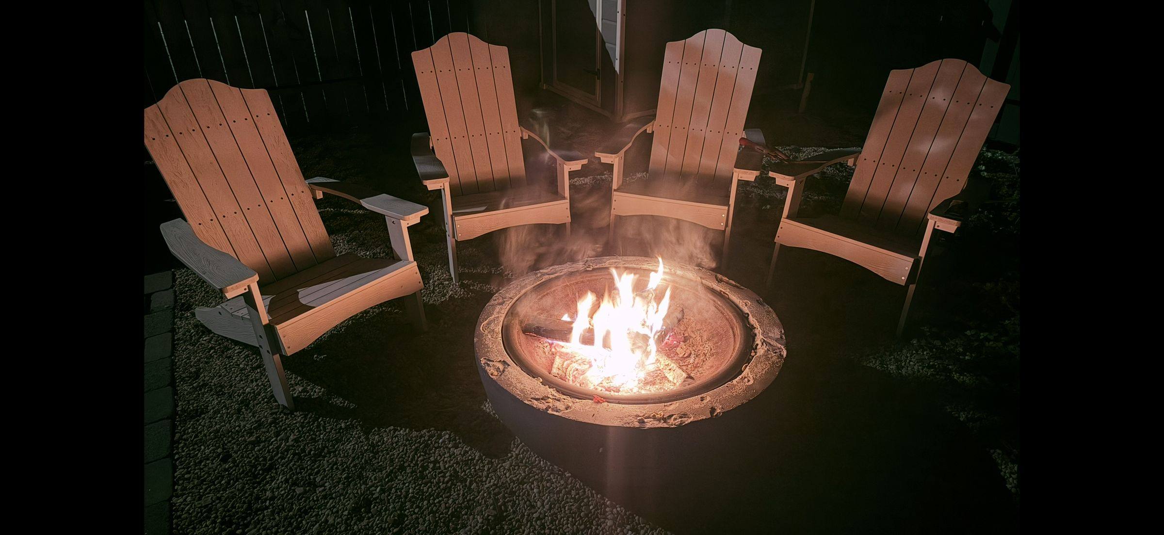Four Adirondack chairs pulled around the backyard fire pit, flames going after dark.