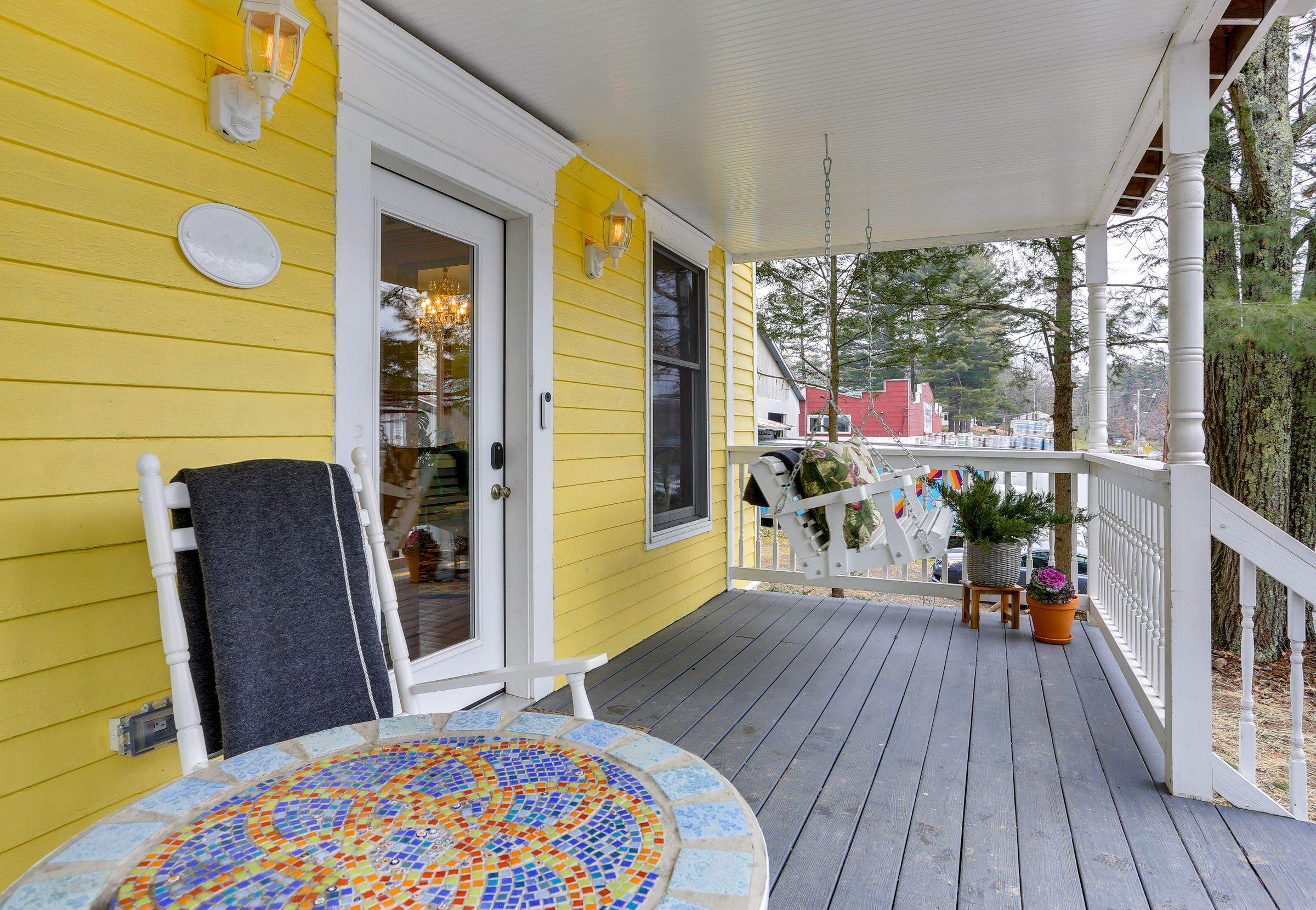The covered front porch of the yellow Victorian with a rocking chair, mosaic table and porch swing.