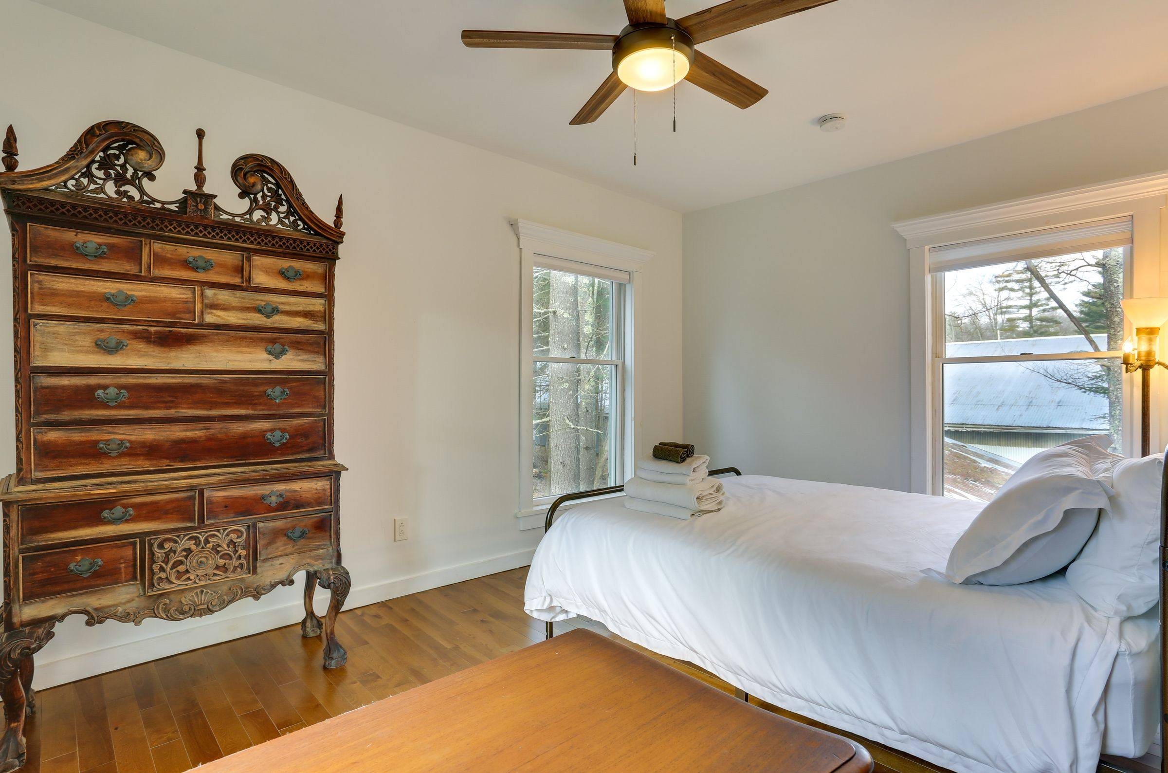 A bedroom corner with an antique carved highboy dresser beside tall windows.