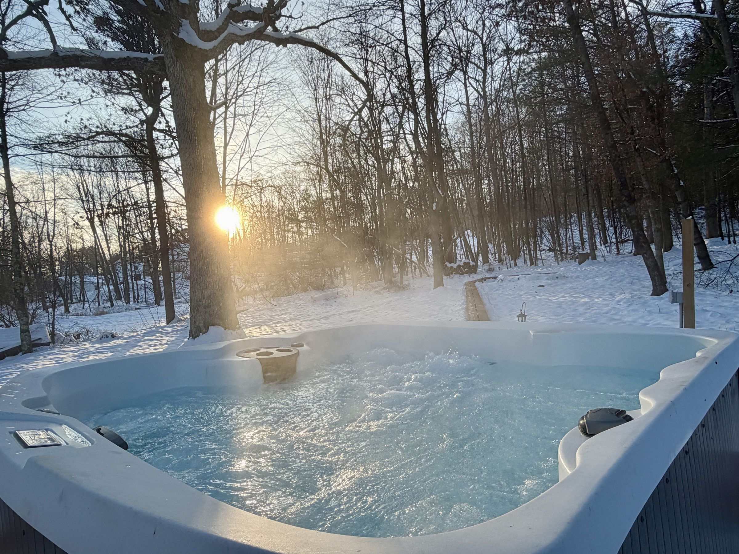 The hot tub steaming at sunset with snow and bare trees in the backyard.